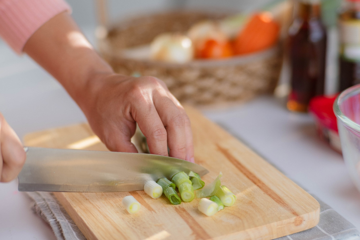 Female hand using a knife to cut Negi (Japanese Long Onion) or Japanese Scallion or Green Onion on wooden cutting board. Woman preparing food in the kitchen at home.